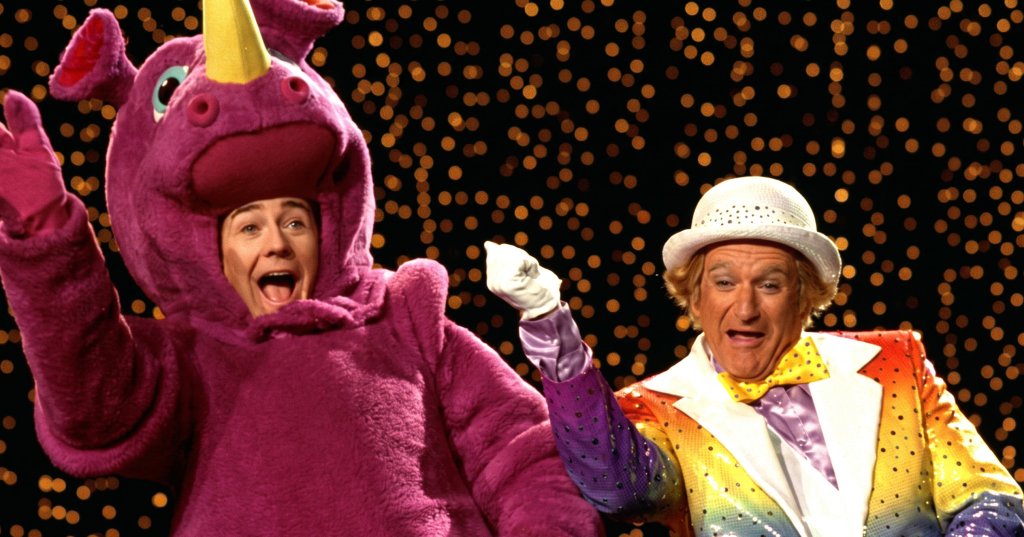 Person in purple unicorn costume with open mouth next to man in sparkly rainbow jacket and white hat against a bokeh light backdrop.