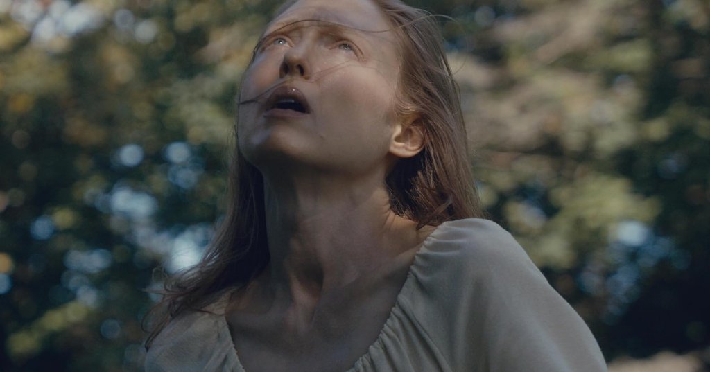A pale woman with long hair tilts her head back and looks up with a horrified expression, outdoors among blurred trees, in a still from Blood Shine (2025).
