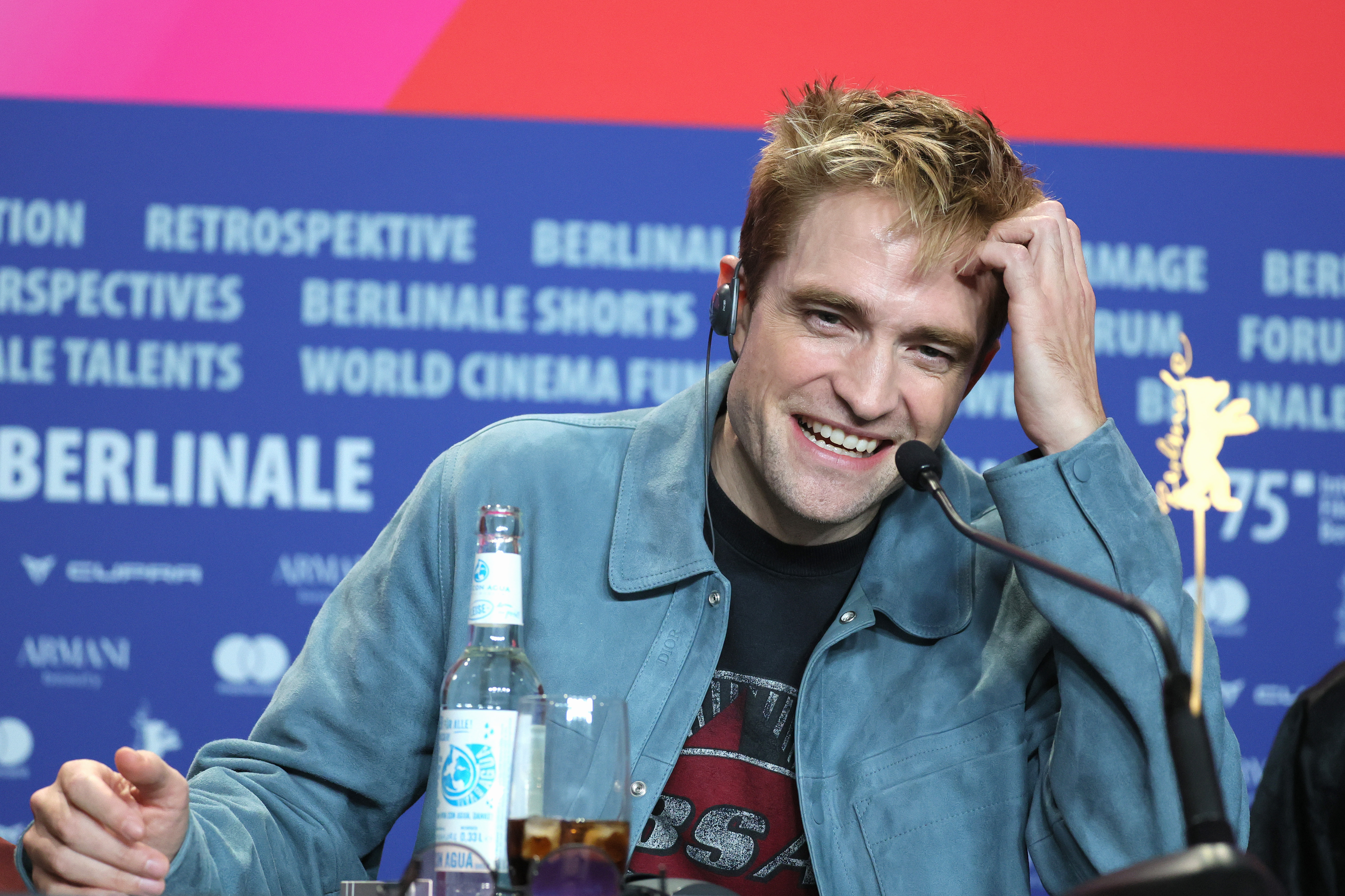 Click to open the image gallery Man in blue-grey jacket laughing at a press conference table with a microphone, water bottle, and a Golden Bear award in front of a Berlinale banner.