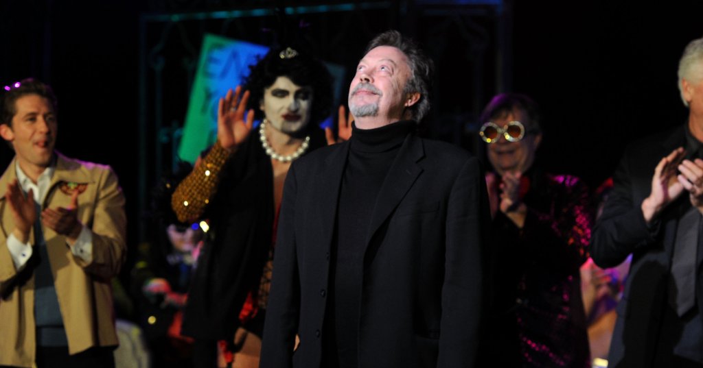 Tim Curry in a black turtleneck and blazer looks upward onstage, surrounded by costumed performers and applauding guests at a theatrical event.