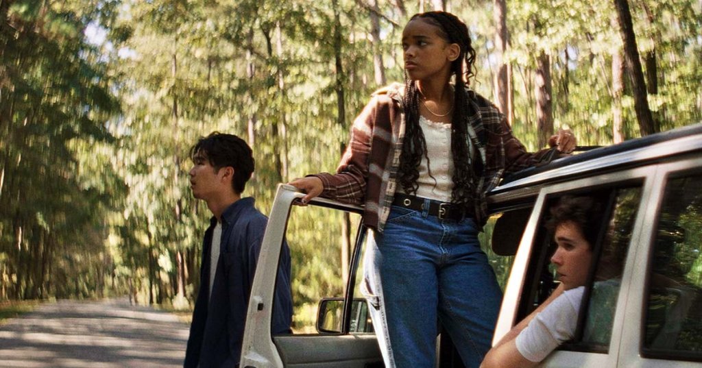 Three teens beside a white van on a tree-lined road; a girl in a plaid shirt stands on the door frame while two boys look away warily.