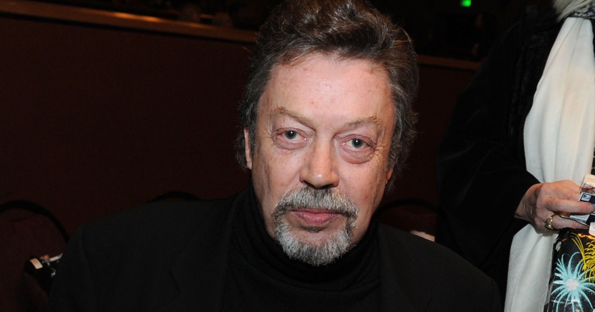 Tim Curry in a black turtleneck and blazer seated at a red velvet-draped table at an indoor event.