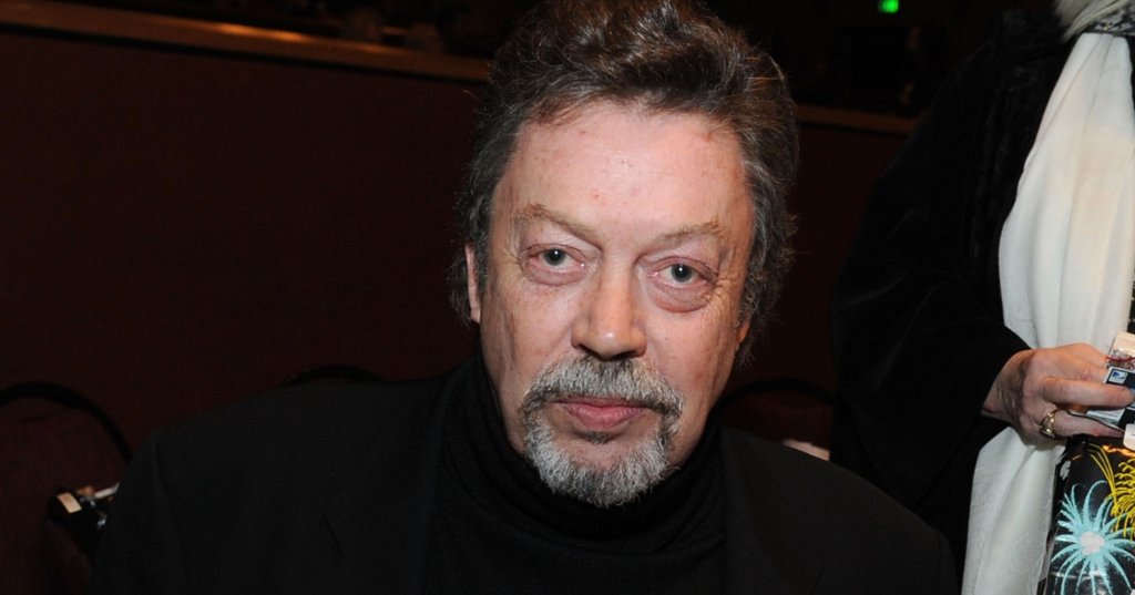 Tim Curry in a black turtleneck and blazer seated at a red velvet-draped table at an indoor event.