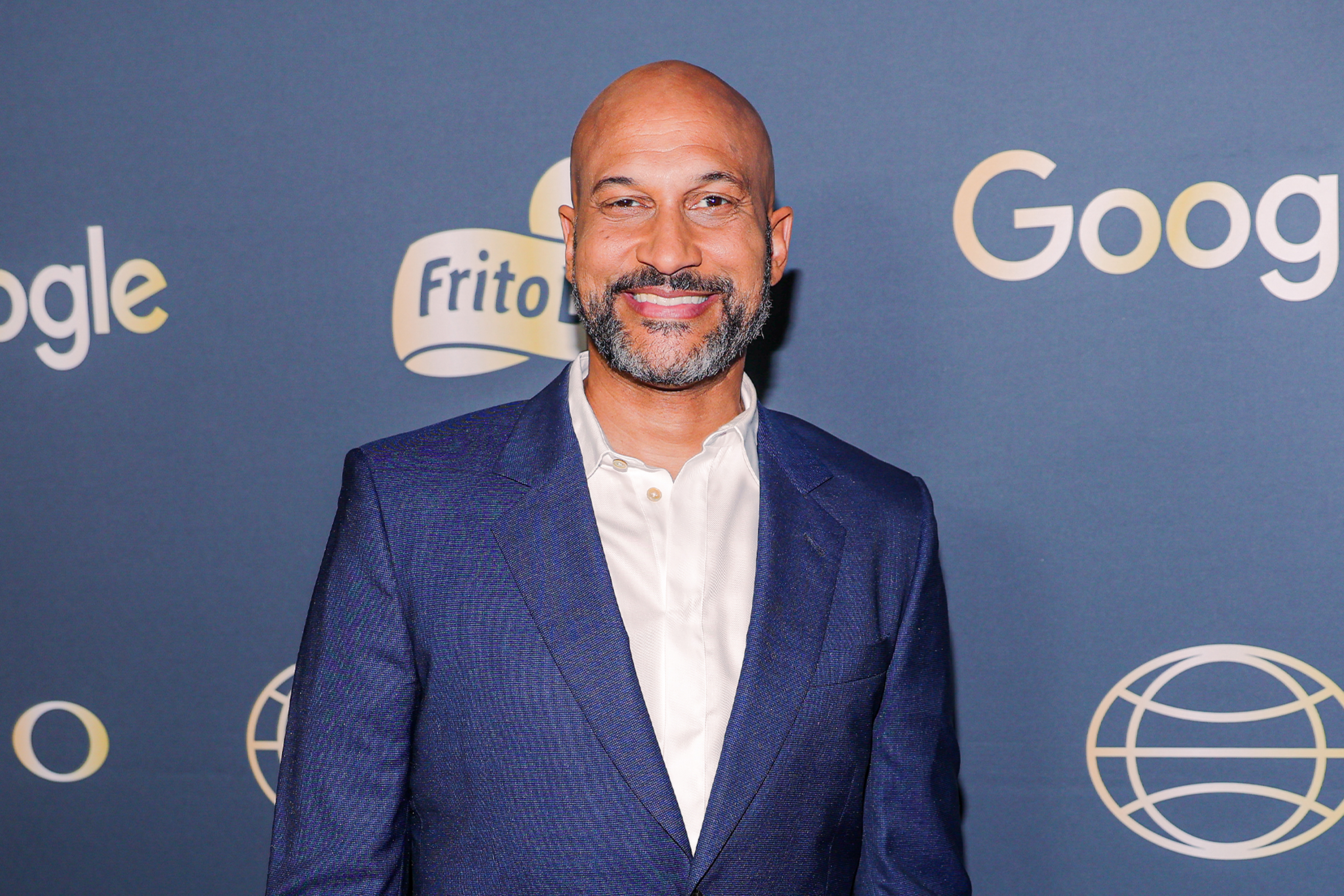 Bald man with a gray beard in a blue blazer and white shirt smiling at a red carpet event with Google and Frito-Lay branding behind him.