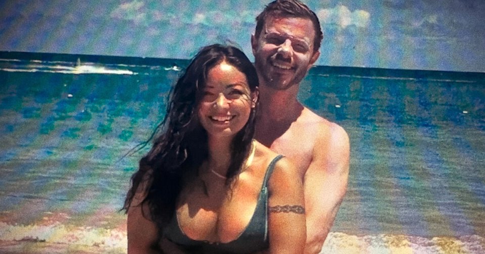 A smiling woman in a bikini and a shirtless man with a tattoo on his arm embrace on a beach with ocean waves behind them.
