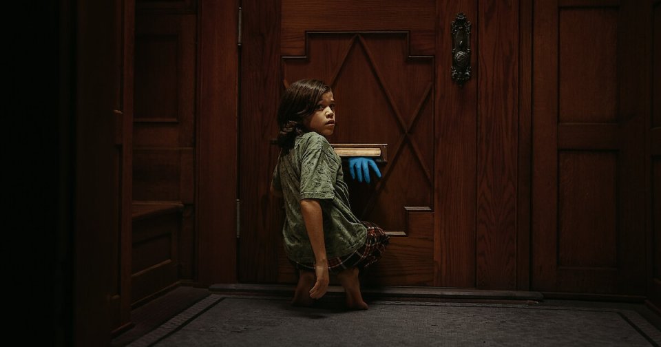 Young barefoot child in a green dress crouching at a dark wooden door while a blue-gloved hand reaches through the mail slot.
