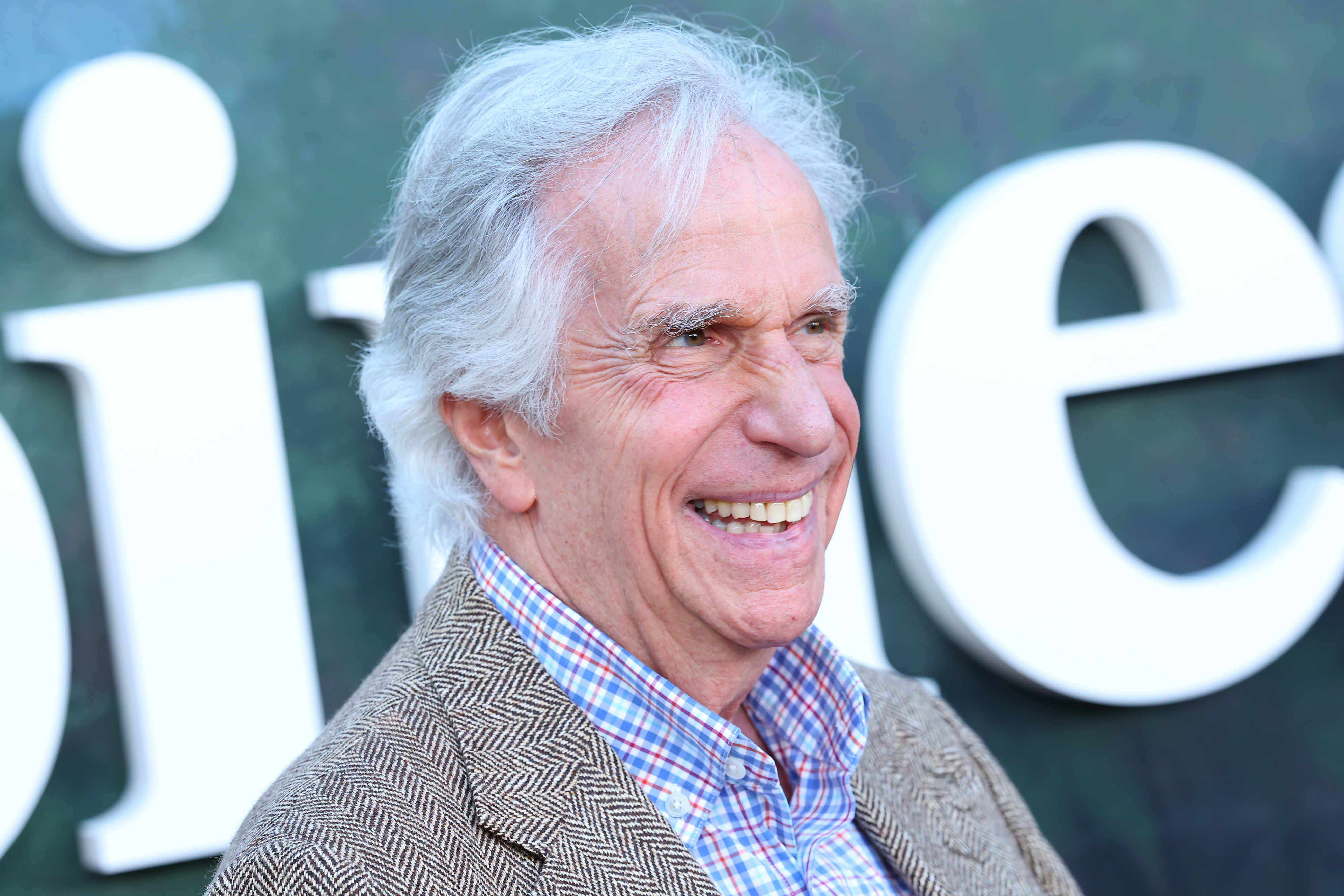 Henry Winkler smiling at a premiere event, wearing a plaid shirt and herringbone blazer, with a teal step-and-repeat backdrop behind him.