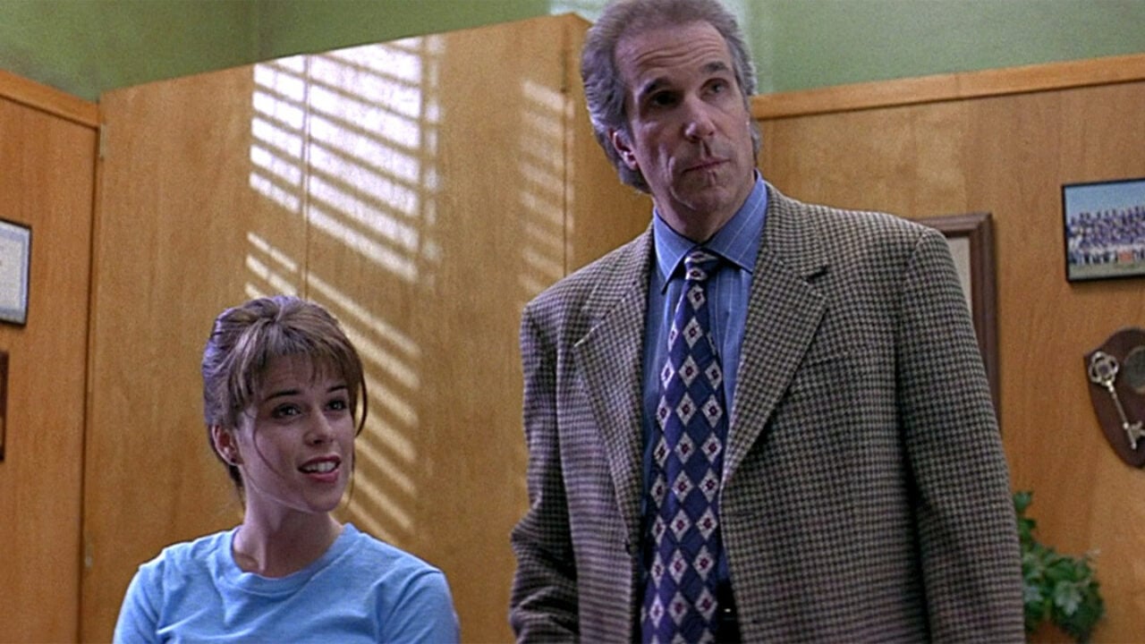A tall man in a houndstooth blazer and patterned tie stands next to a shorter smiling young woman in a blue shirt inside a wood-paneled school office.