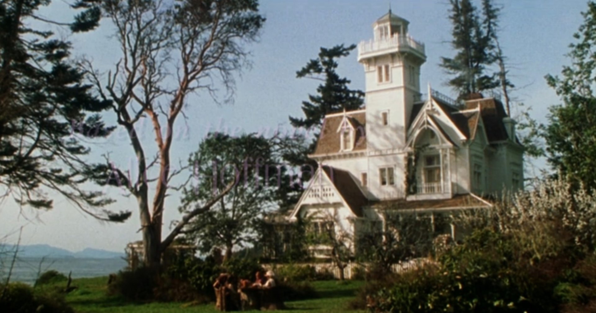 Wide exterior shot of a large white Victorian mansion with a tall tower, surrounded by trees, with mountains visible in the background.